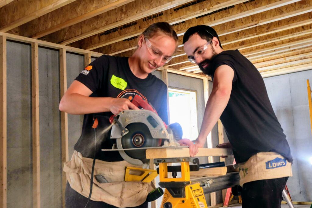 Tiffany and Trevor in the Habitat for Humanity house's basement, working together to cut a board with a circular saw.
