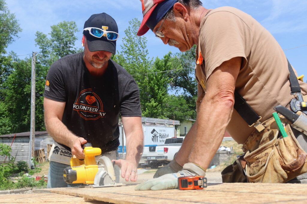 Ryan working with a Habitat for Humanity volunteer leader to cut a piece of manufactured wood with a circular saw outside the house.